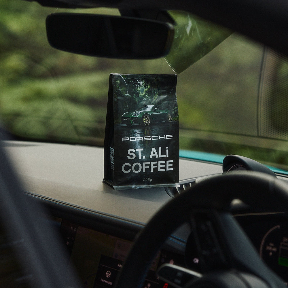 A bag of St. Ali Coffee by Porsche is displayed on a car dashboard, framed by the steering wheel and windshield with a blurred outdoor background.